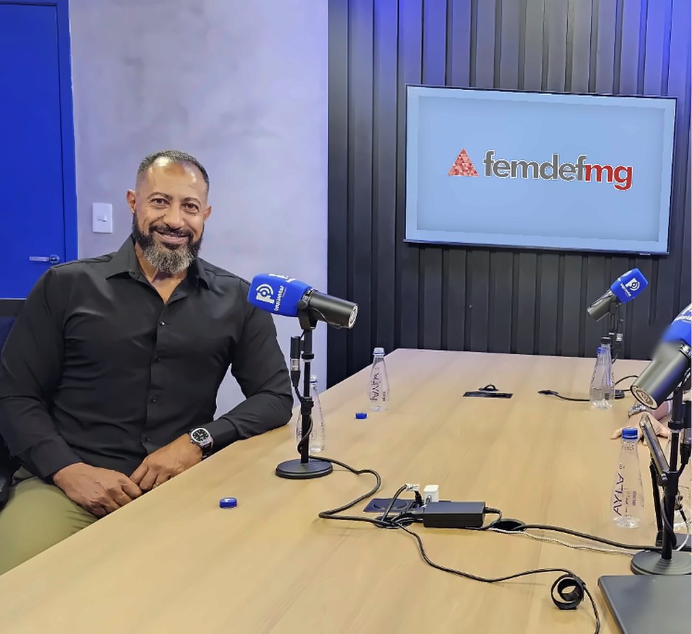 Wellington Detroit, presidente da FEMDEFMG, sorrindo em um estúdio com a logo da federação na tela ao fundo
